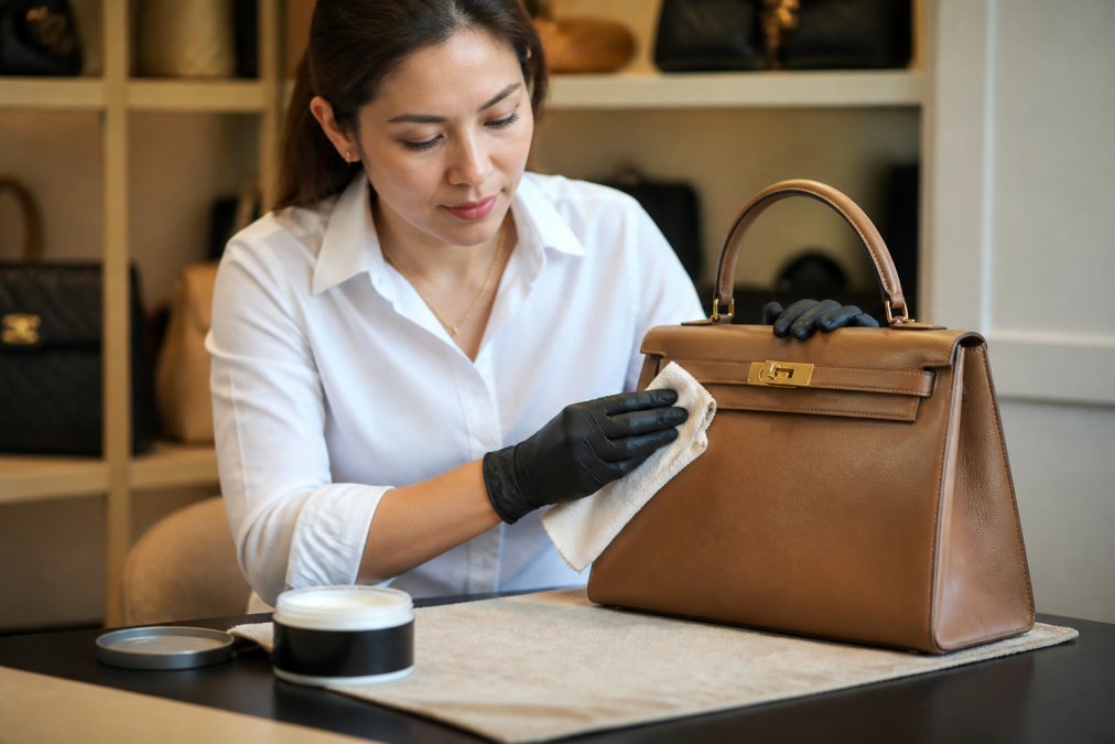 Hands carefully applying leather conditioning cream to a structured handbag with a soft cloth, demonstrating proper maintenance technique on a clean work surface