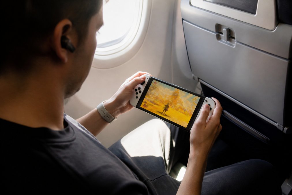 Traveler in airplane window seat holding Nintendo Switch OLED with vibrant game display visible, natural sunlight streaming through window, compact console fitting easily in lap