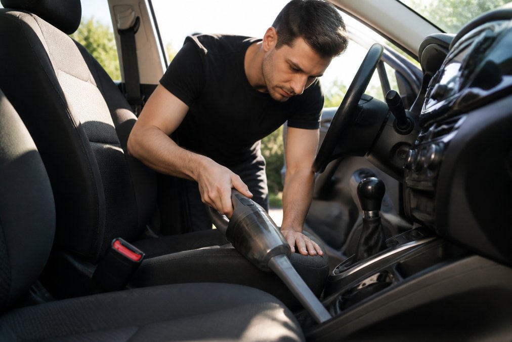 Person using a handheld vacuum with a crevice tool to clean dirt from a car seat gap and floor mats.