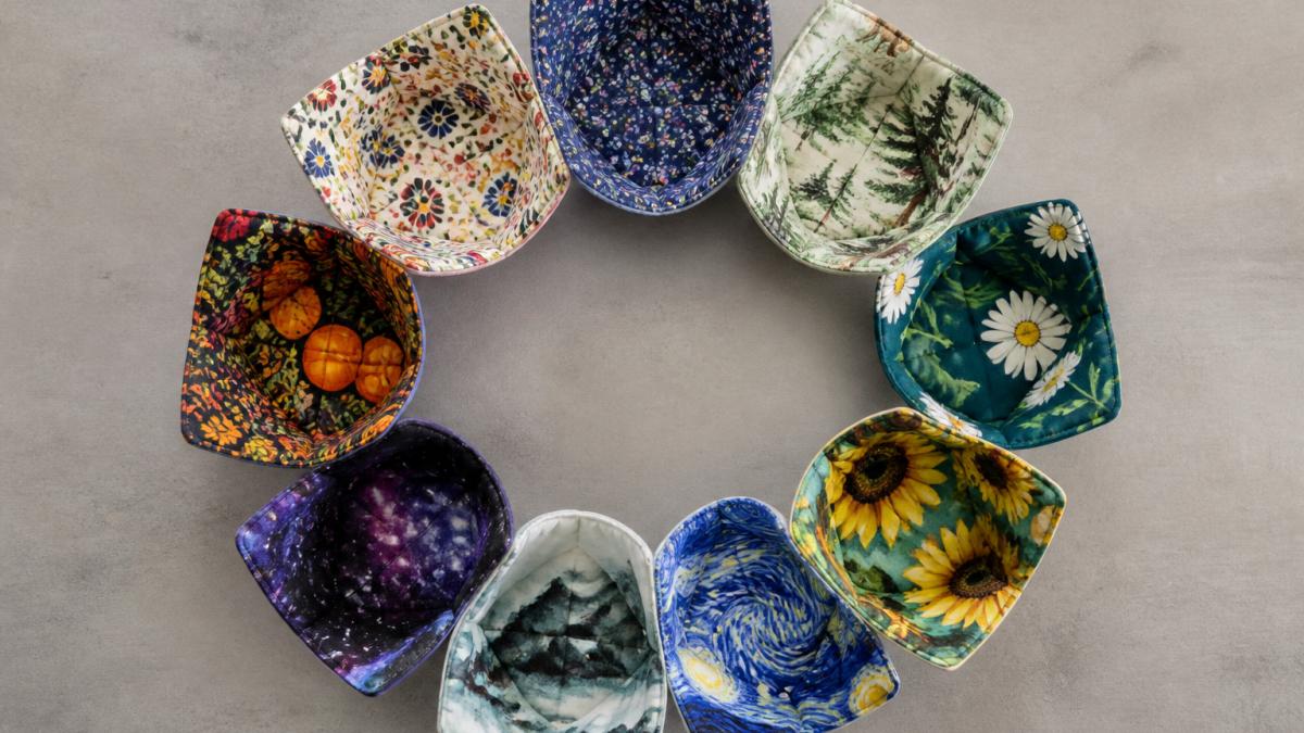 Flat lay of colorful handmade bowl cozies arranged in a circle on a neutral background, showcasing multiple designs including floral patterns, nature themes, and artistic illustrations in vibrant colors