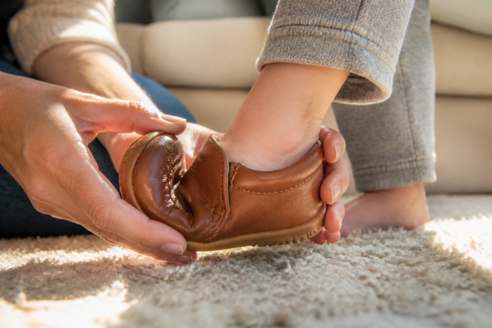 Close-up of a toddler's small foot being gently fitted into a soft leather shoe, showing the flexible sole bending naturally and padded collar supporting the ankle, with natural daylight highlighting the quality craftsmanship
