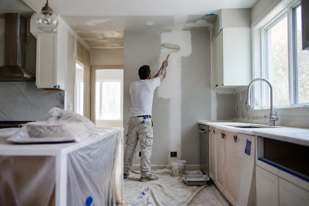 Skilled tradesperson performing drywall repair and painting in residential kitchen with professional equipment and drop cloths, natural daylight from windows