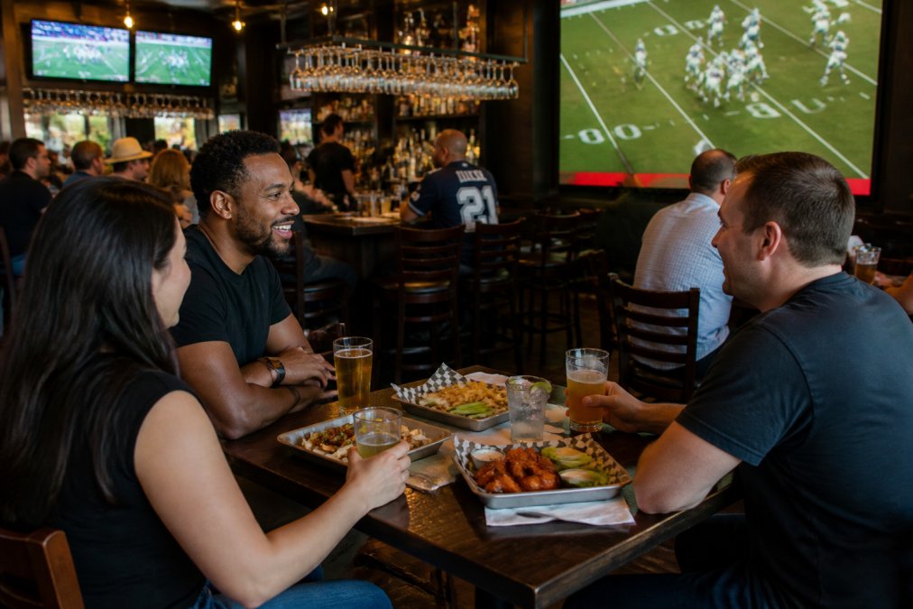 A lively sports bar interior with patrons watching a game on large screens, enjoying wings and drinks at high-top tables under warm ambient lighting