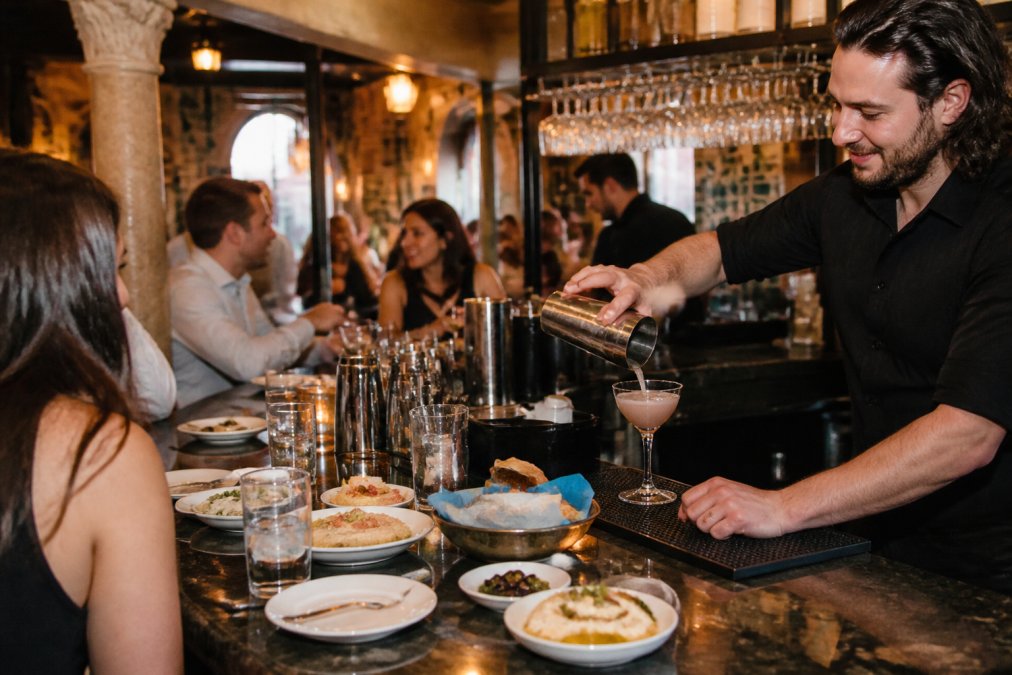 A lively bar area at a Greek restaurant with a bartender pouring a cocktail, small plates of mezze on the counter, and a few patrons chatting in the background