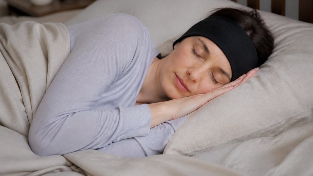 Person lying on their side in bed wearing soft headband headphones, relaxed expression, natural bedroom lighting with pillows and neutral bedding