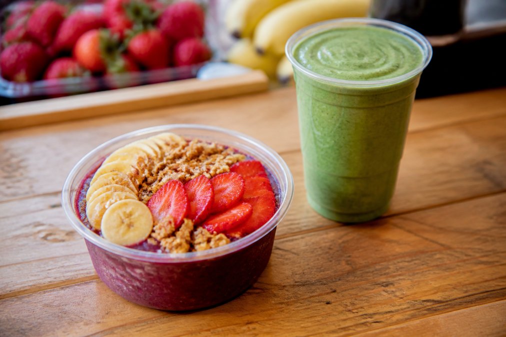 A close-up of a freshly blended açaí bowl topped with granola, banana slices, and strawberries on a wooden counter, with a green smoothie beside it in a clear cup