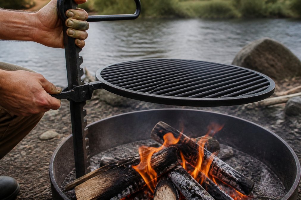 Hands adjusting the locking mechanism to raise a heavy carbon steel grill grate over an active campfire, flames visible below
