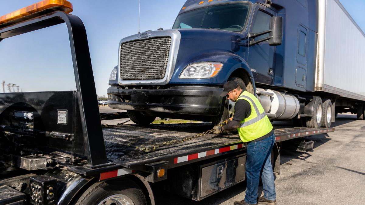 Professional heavy-duty flatbed tow truck with operator securing a large commercial semi-truck during daytime recovery operation, showing proper equipment and rigging setup