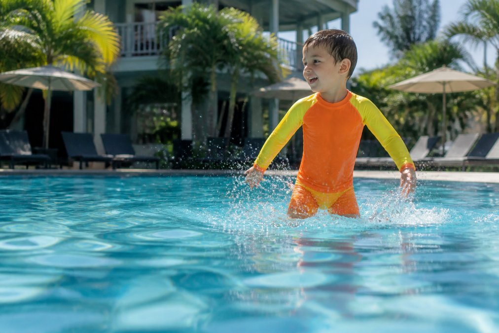 A young boy wearing a neon orange and yellow long sleeve rash guard set splashing in a sunlit outdoor pool, viewed from across the water at water level