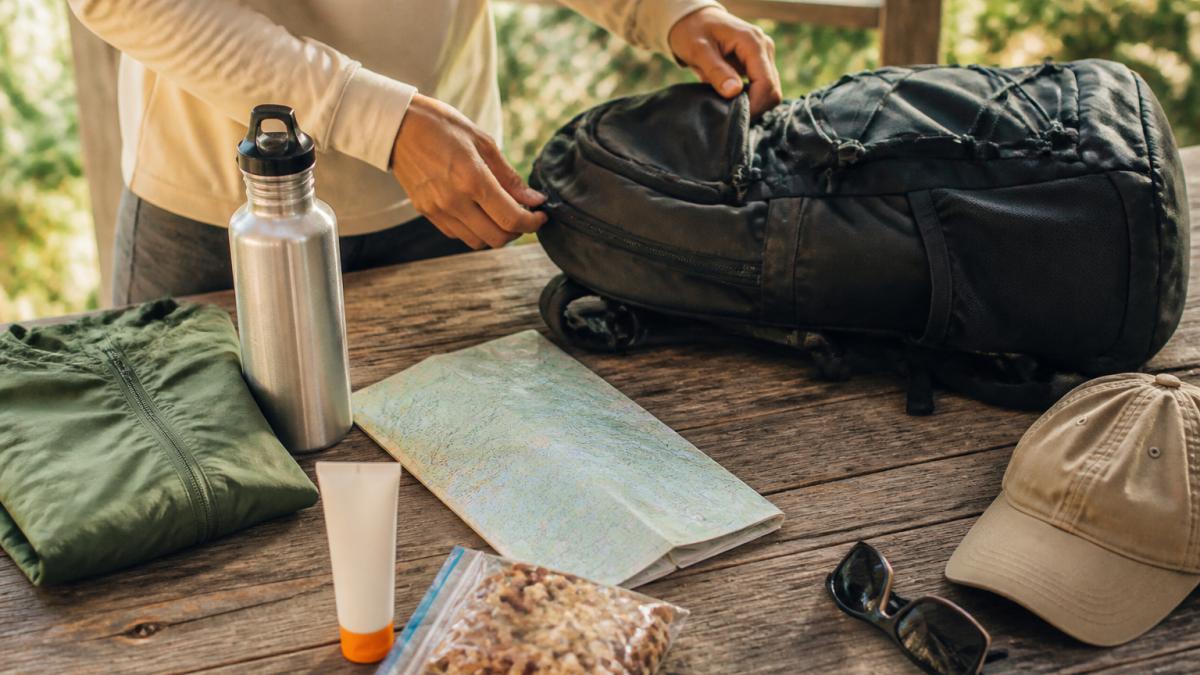 A beginner hiker laying out day-hike essentials on a wooden table, including a water bottle, folded rain jacket, sunscreen, snacks, a paper map and a small backpack, in soft morning light