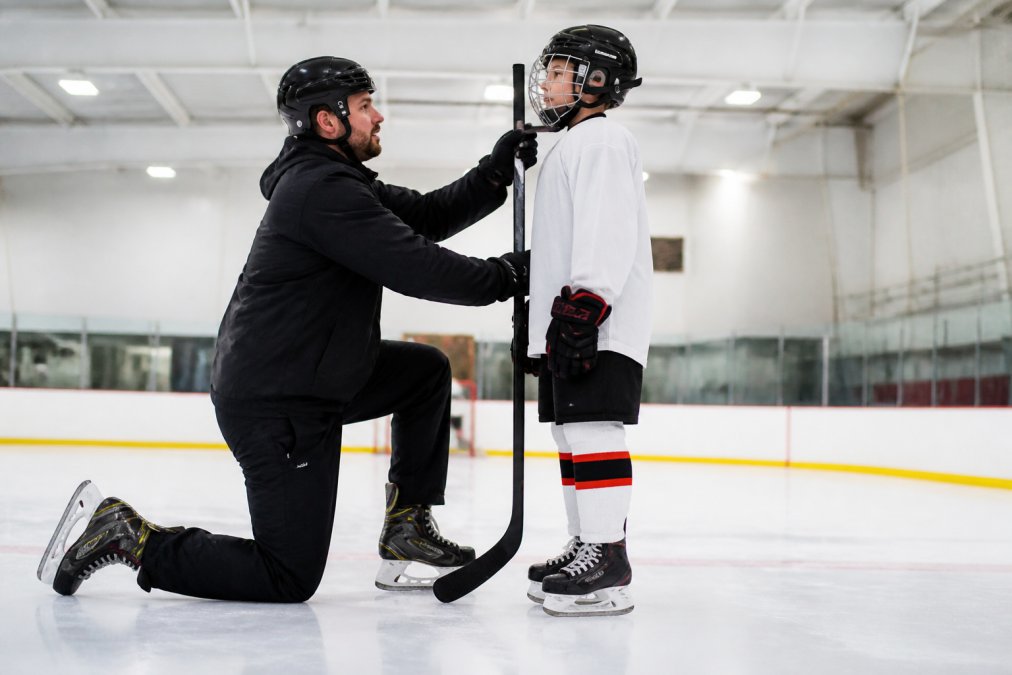 A young hockey player in full gear standing next to a coach in a bright indoor arena, holding a stick upright against their body while the coach checks the length against the child's chin