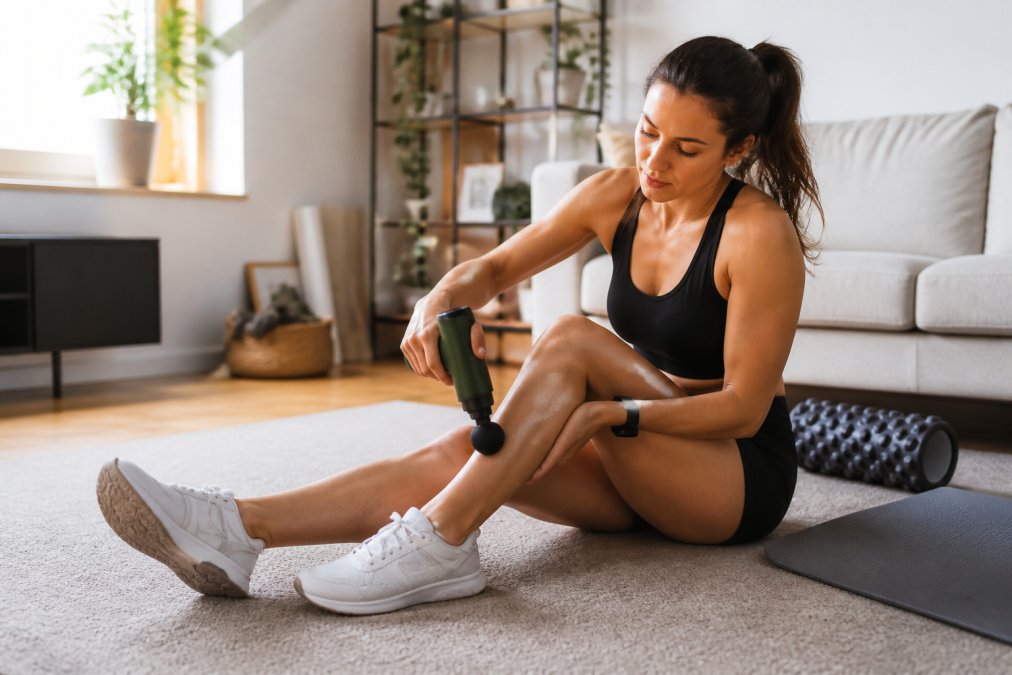 A woman in athletic wear sitting on a living room floor using a green massage gun on her calf muscle, with a foam roller and yoga mat nearby in a bright home gym setting