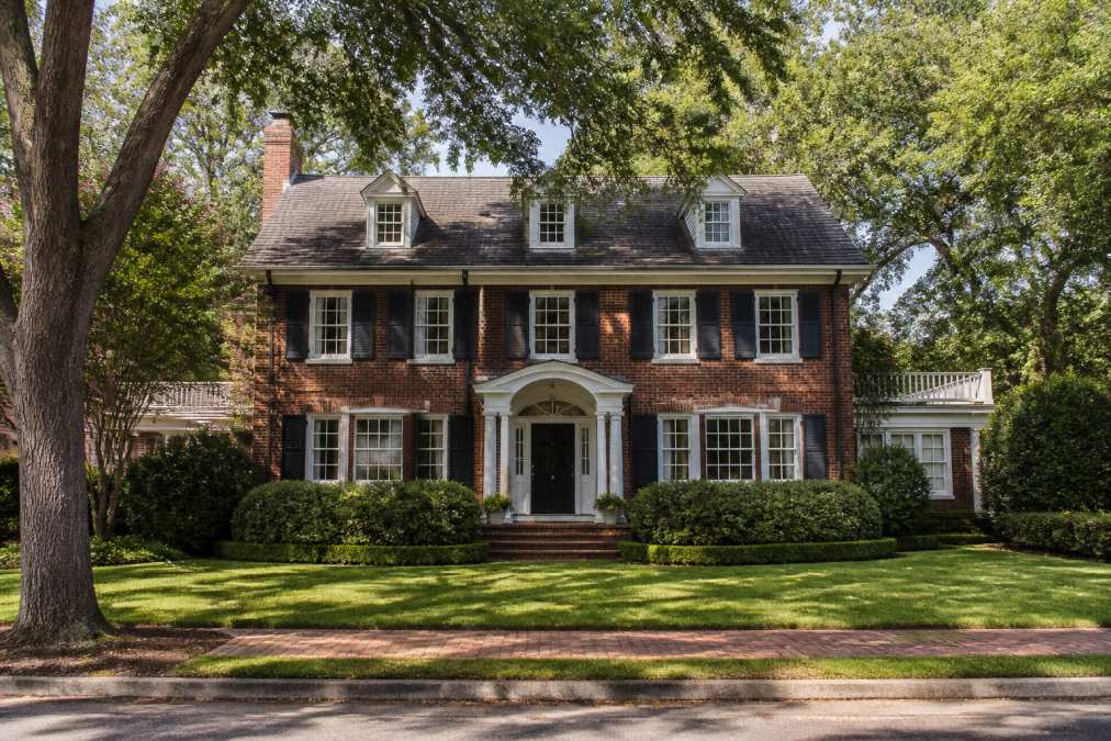 Exterior of a beautifully maintained 1920s colonial home with original brick facade, white trim, multipane windows, and mature landscaping on a tree-lined street in afternoon sunlight