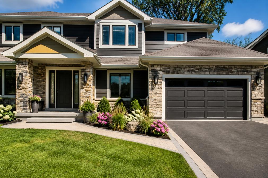 Modern residential home exterior featuring a gleaming steel entry door with sidelights, manufactured stone veneer accents on the front facade, freshly painted trim, manicured landscaping with flowering shrubs, and a newly replaced garage door under bright afternoon sunlight