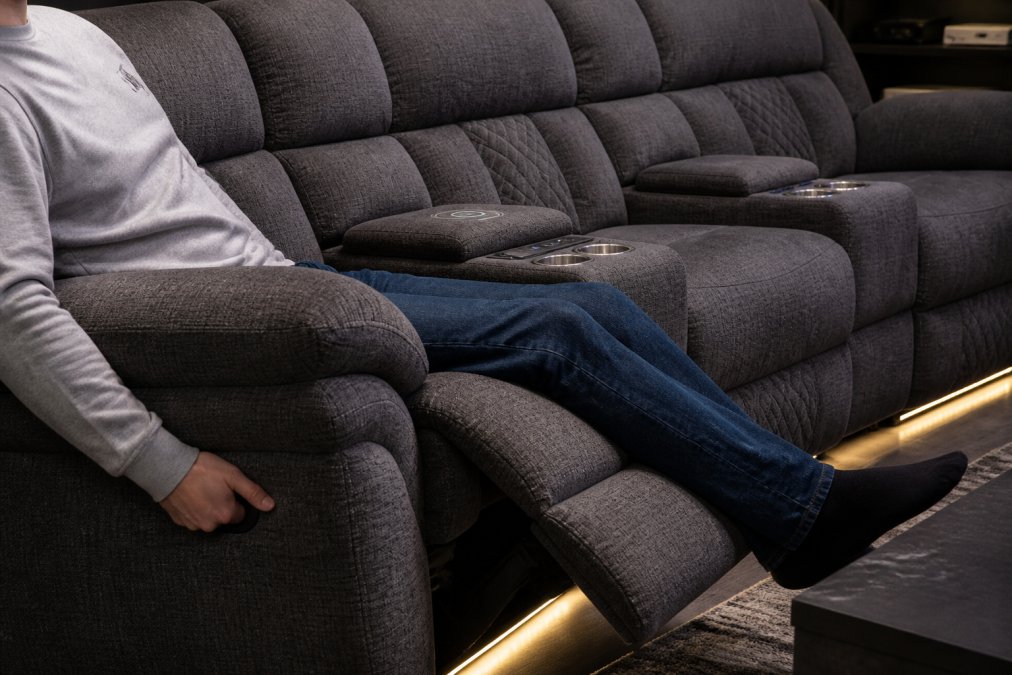 Close-up of luxury power reclining sofa with diamond stitching detail, showing the wireless charging center console with cup holders, someone's hand using the power recline control, warm LED accent lighting visible beneath the seat