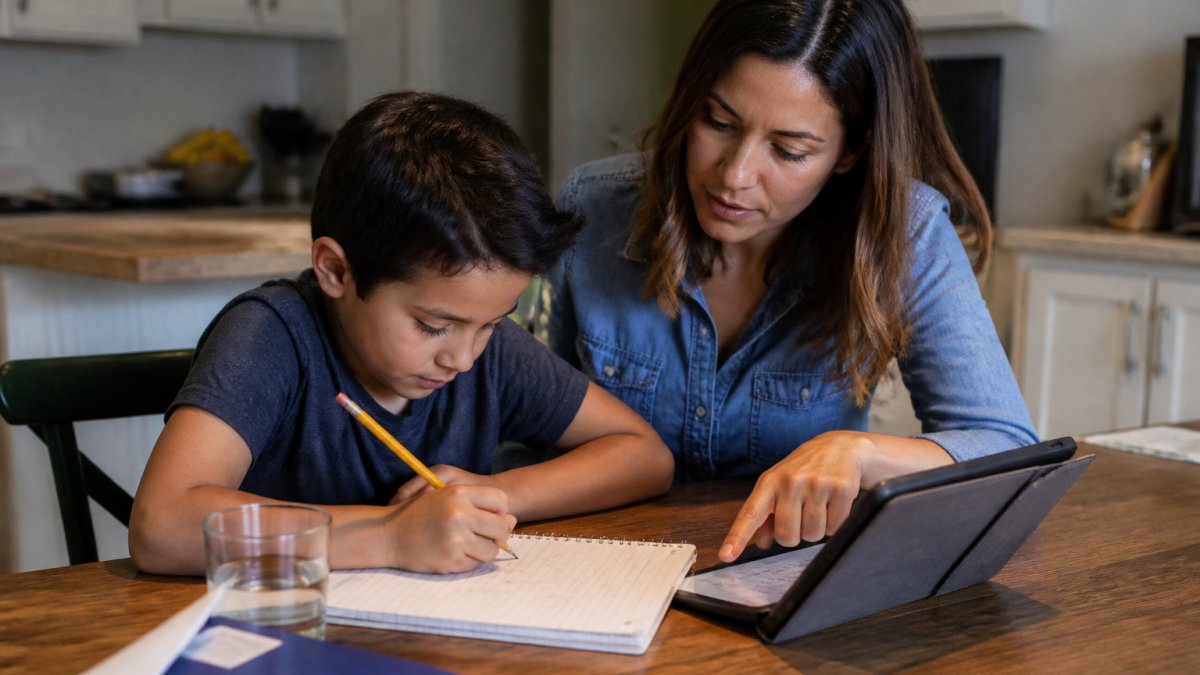 A parent and elementary-aged child sitting together at a kitchen table in the evening, child writing in a notebook while the parent points at a tablet screen showing a homework assignment