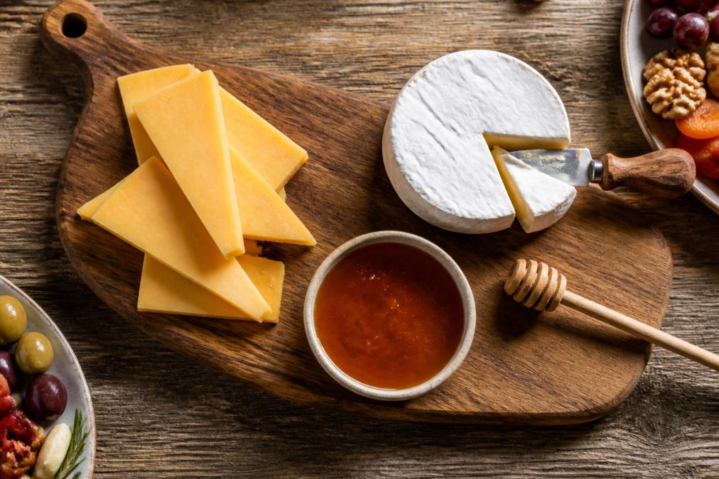 Overhead shot of a rustic wooden charcuterie board with slices of aged cheddar, a wheel of creamy brie with a knife, and a small ceramic dish of hot honey with a wooden honey dipper beside it, soft natural window light