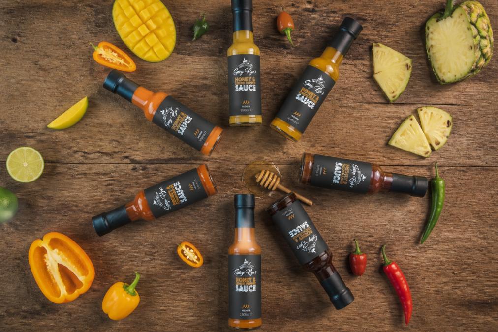 Overhead flat-lay of seven sauce bottles arranged in a circle on a wooden surface, showing distinct colours from bright lemon yellow through mango orange to deep smoky brown, with fresh ingredients scattered between them including whole peppers, mango slices, honey drizzle, and pineapple chunks, natural window lighting