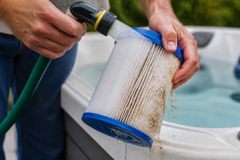 Close-up of hands rinsing a hot tub cartridge filter under running water from a garden hose, showing debris flowing away from the pleated filter surface in natural daylight