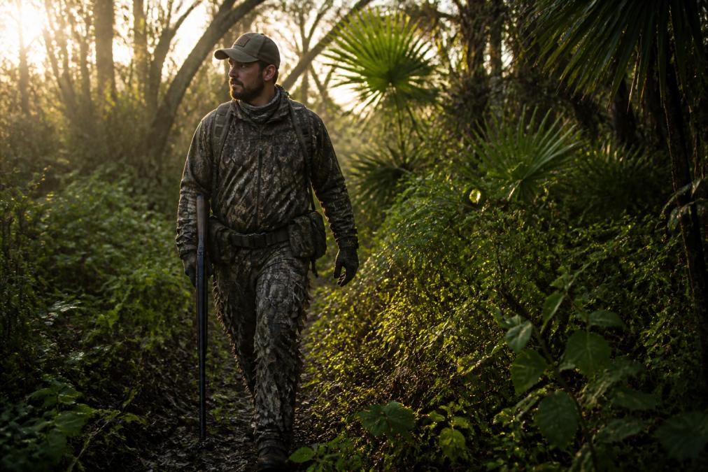 A hunter in durable camouflage clothing and brush pants walking through thick vegetation with a rifle, early morning light filtering through trees