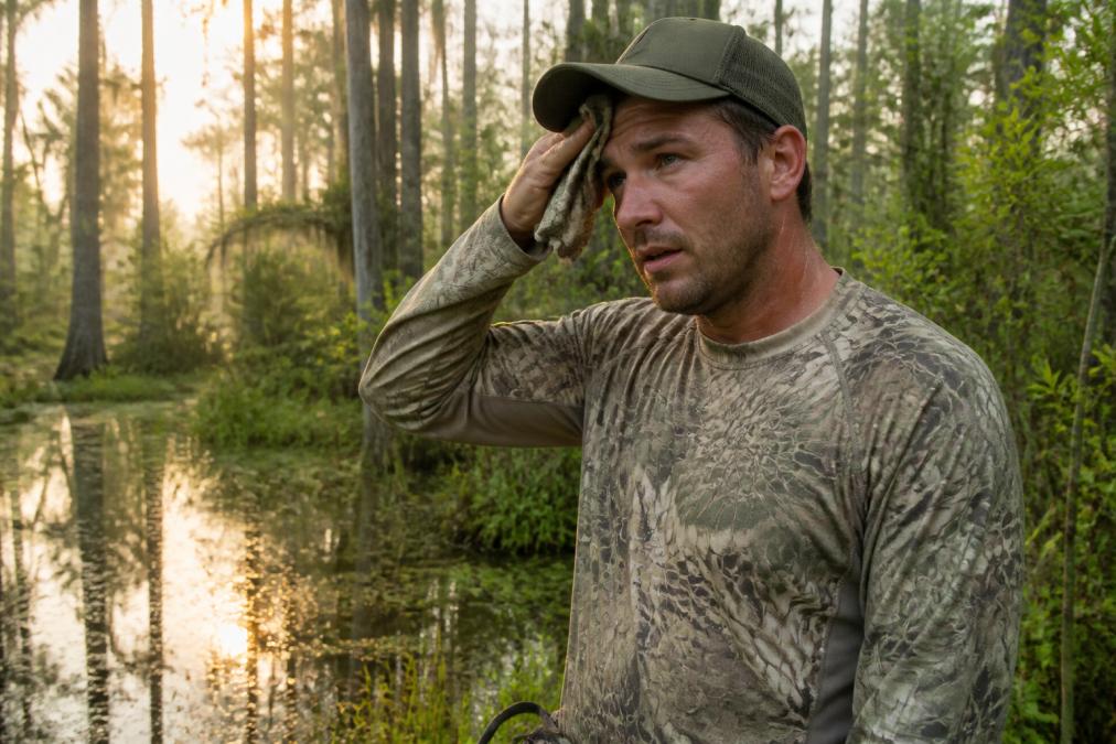 A hunter in a moisture-wicking shirt and low-profile cap standing in a humid cypress swamp during early morning light, wiping sweat from forehead, with shallow water and vegetation visible in the background