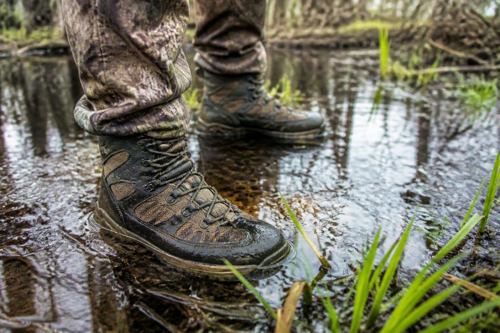 A close-up of a hunter's feet in breathable boots standing in shallow swamp water, showing mesh venting and water-resistant material with muddy terrain and vegetation visible