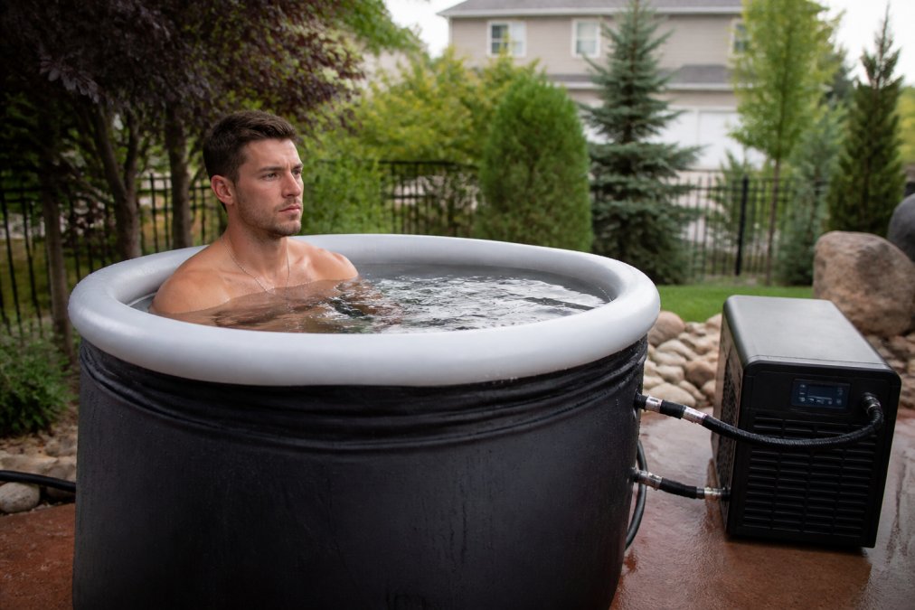 An athlete in athletic wear sitting inside a large portable inflatable ice bath tub outdoors, water at chest level, with a focused expression and a water chiller unit visible beside the tub