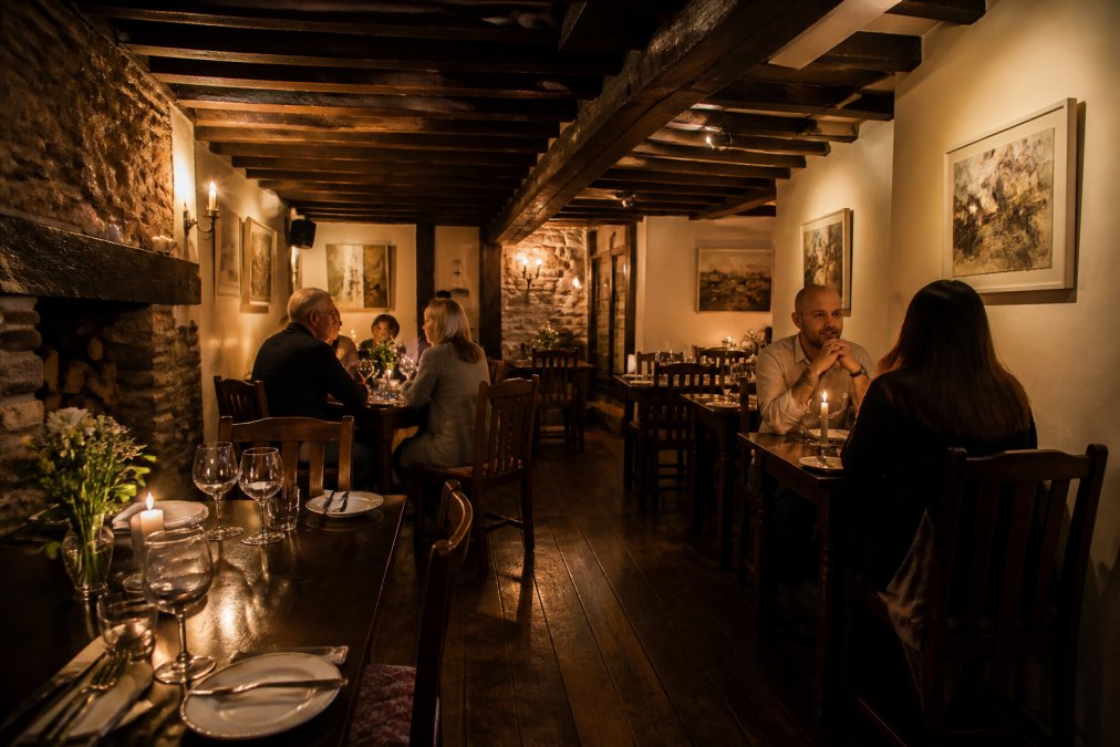 A cosy independent restaurant interior with historic wooden beams and exposed stonework, candlelit tables arranged intimately, local artwork hanging on cream-coloured walls, warm ambient lighting creating shadows across the intimate dining space, and a few diners seated at tables in the background