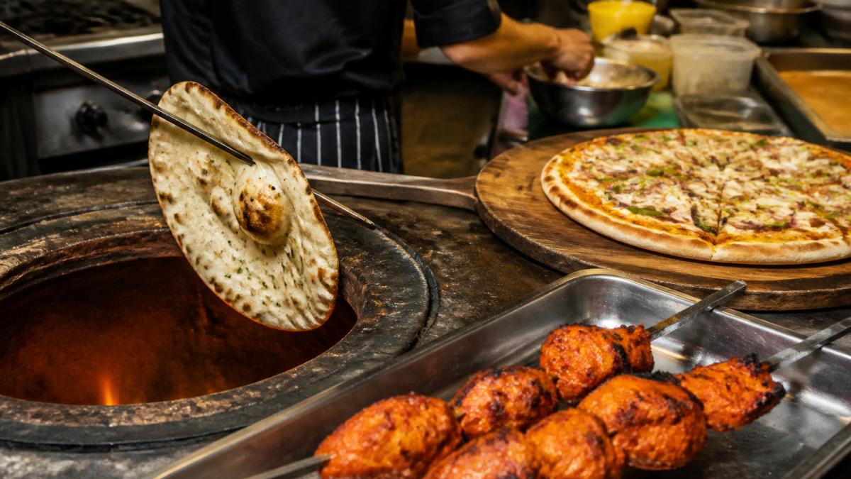 A freshly baked naan bread being lifted from a rotary tandoor with flames visible, alongside a tray of chicken tikka skewers and a handmade pizza on a wooden board in a busy takeaway kitchen