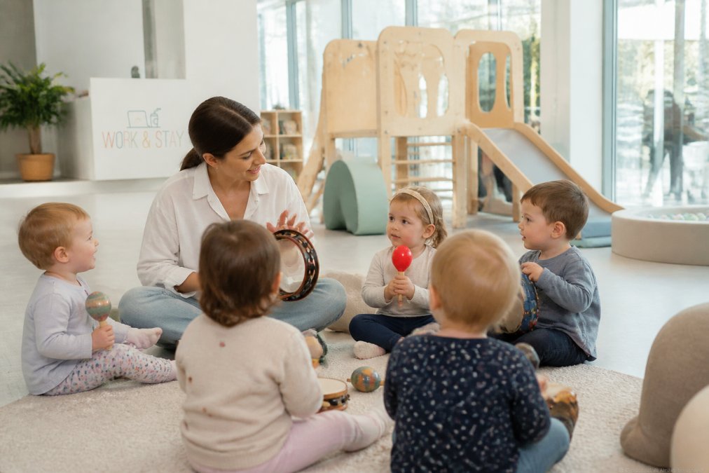 Toddlers and infants engaged in guided music activity with an instructor playing instruments, colorful play equipment visible in background, bright natural lighting through large windows in modern facility