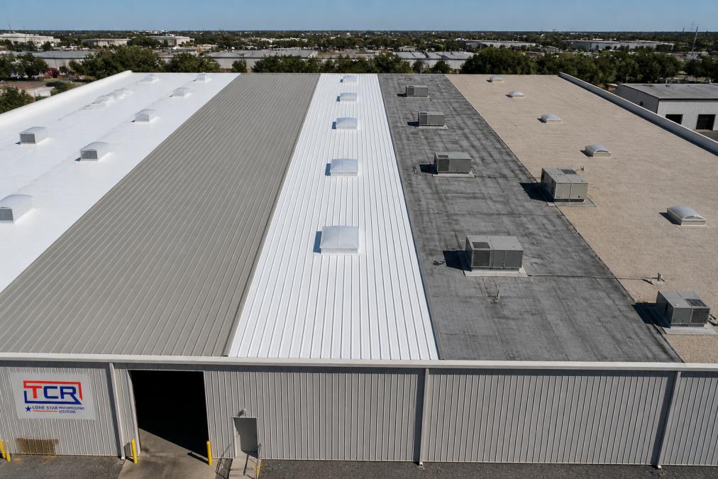 Overhead view of a large industrial warehouse roof showing different roofing material sections under bright Texas sunlight, with heat shimmer visible on metal surfaces