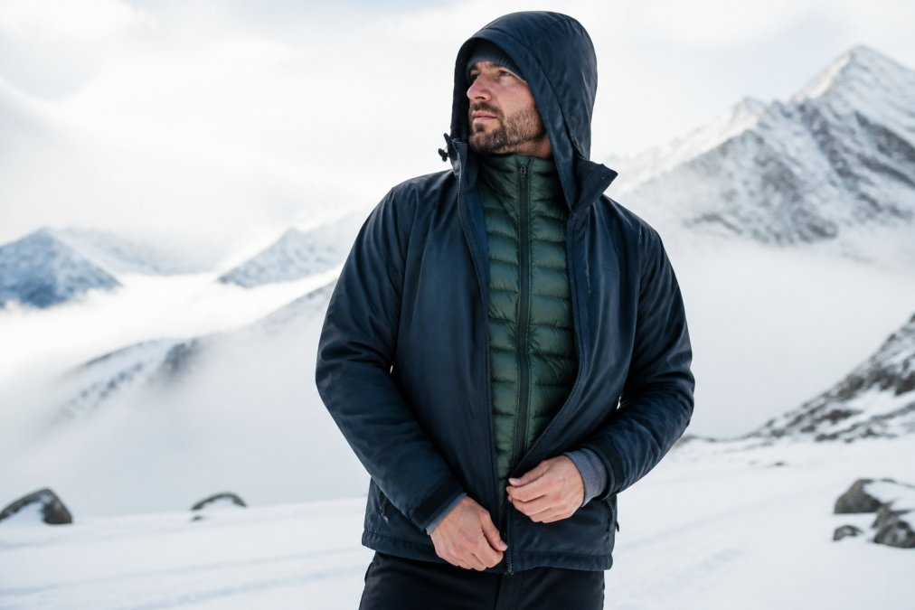 Outdoor enthusiast wearing complete winter layering system, visible base layer at wrists, insulated puffer jacket as mid-layer, and waterproof outer shell jacket, standing in snowy mountain landscape with misty conditions