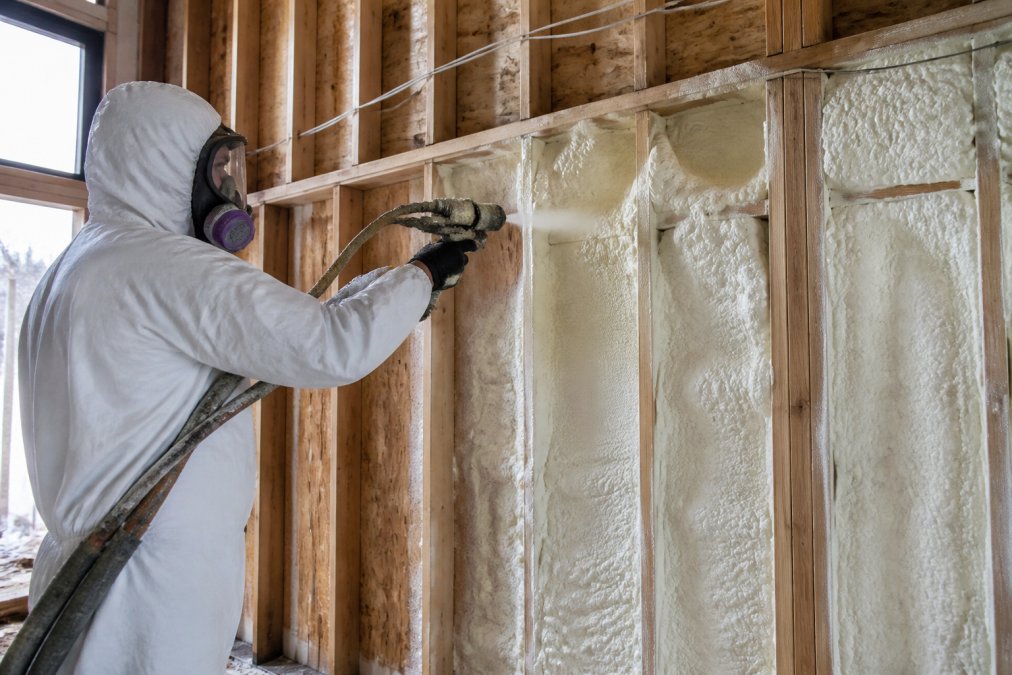Professional contractor applying closed-cell spray polyurethane foam to wall cavities and rim joist with spray gun, showing foam expansion and coverage in a home addition frame during cold season