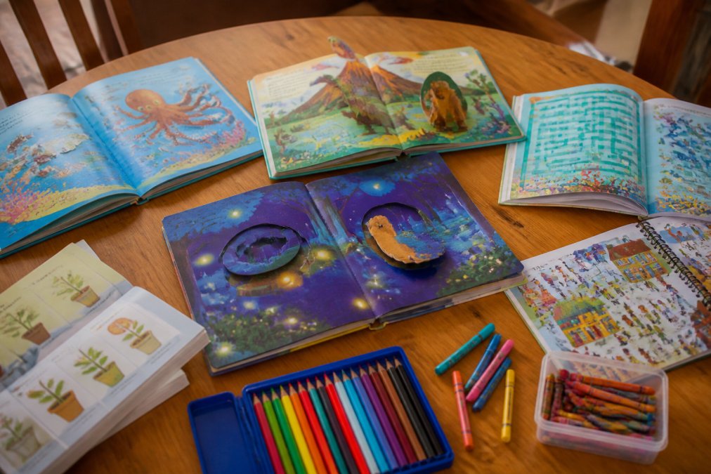 Collection of colorful storybooks and activity books spread across a wooden table with crayons, markers, and colored pencils nearby, showing the variety of interactive options available to young readers