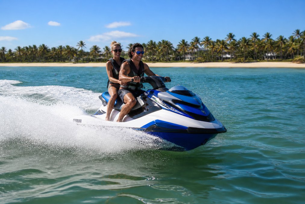 Two people riding Yamaha personal watercraft at speed on calm Gulf Coast water, sandy shoreline and palm trees visible in the background under a bright blue sky, sunlight reflecting off the water