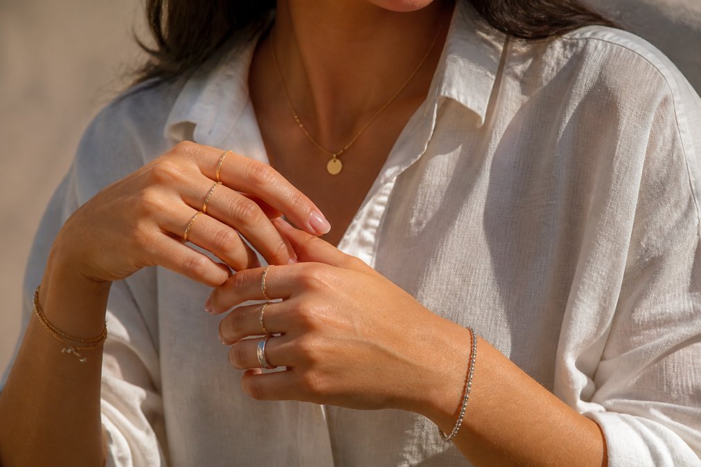 Close-up of a woman's hands and wrists displaying layered gold and silver rings, delicate bracelets, and a minimalist necklace against a neutral background with natural daylight