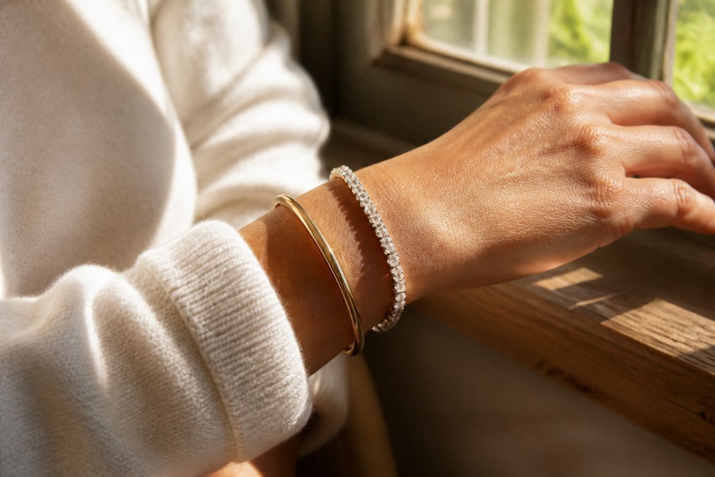Close-up of a woman's wrist wearing a sparkling sterling silver tennis bracelet alongside a simple gold bangle, with natural light streaming through a window highlighting the stones and casting soft shadows on her skin