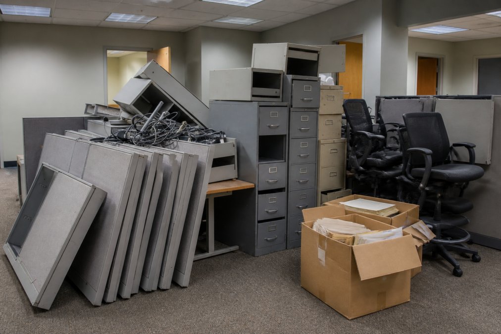 Commercial office space with stacks of metal file cabinets, old fabric cubicle partitions, broken ergonomic chairs, and cardboard boxes of archived documents ready for removal, photographed under fluorescent office lighting