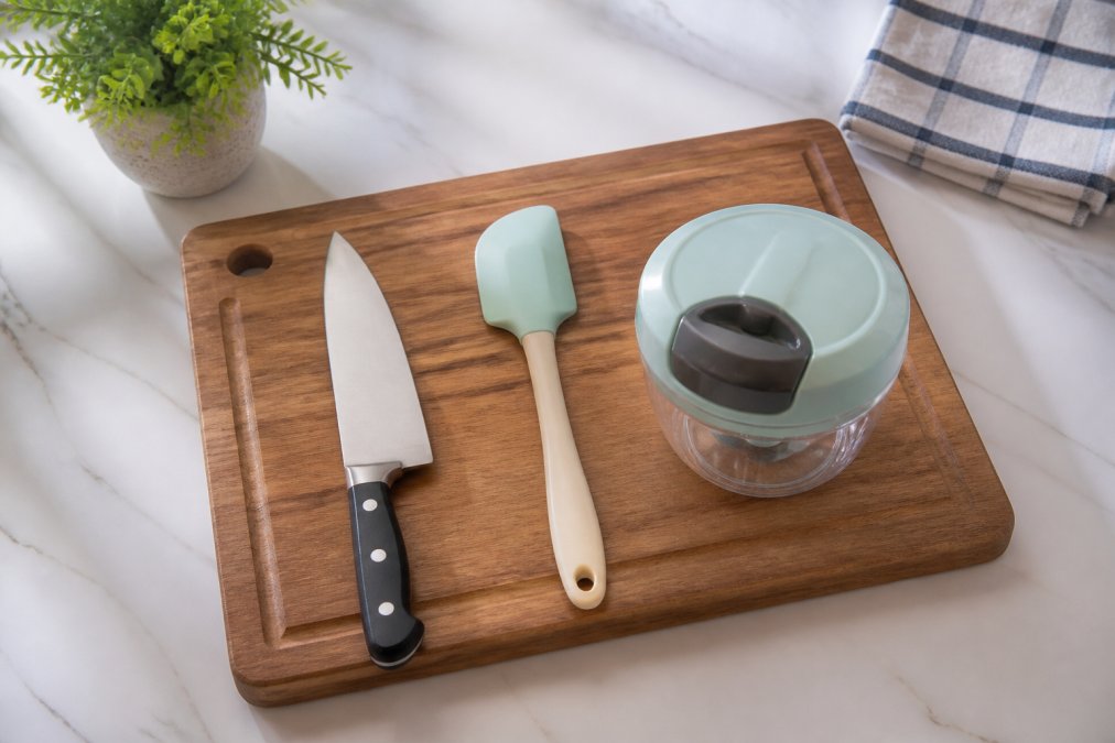 Overhead flat-lay of a wooden cutting board, chef's knife, silicone spatula, and hand chopper arranged neatly on a clean white kitchen counter with natural light