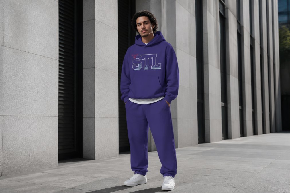 Young professional wearing layered streetwear in urban downtown setting, oversized boxy hoodie in muted tone over a raw-seam tee, relaxed-fit sweatpants, standing against modern architecture with natural daylight
