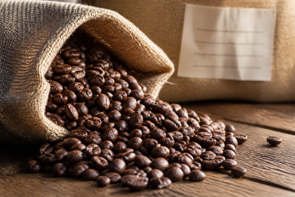 Close-up of freshly roasted whole coffee beans spilling from a burlap sack onto a wooden table, with a small roasting date stamp visible on the bag in the background