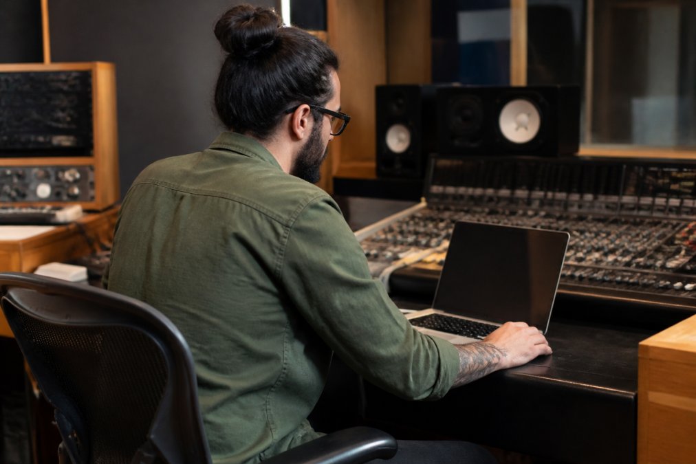 A music label manager reviewing royalty reports on a laptop in a professional recording studio, audio monitors and a mixing console visible in the background