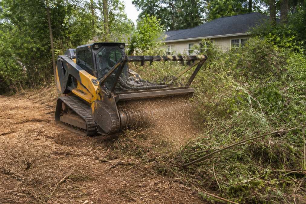 Heavy forestry mulching equipment with rotating grinding head actively processing brush and small trees into mulch on an overgrown residential property, with fine mulch material visible on the ground