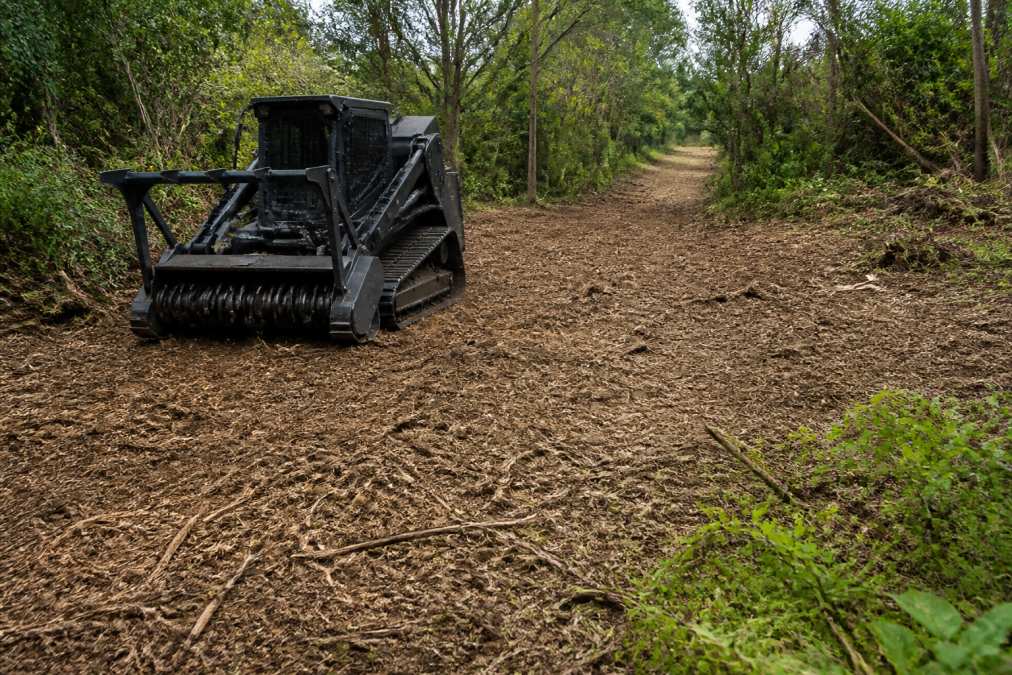 A forestry mulcher clearing dense brush on a hunting ranch, leaving a thin layer of mulch on the ground with green regrowth visible along the edges, deer trail in the distance