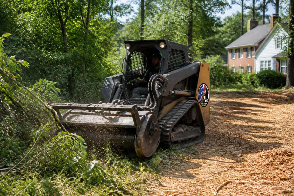 Professional operator in cab of forestry mulcher grinding dense brush and small trees into mulch on residential property, with fresh mulch visible on ground behind equipment