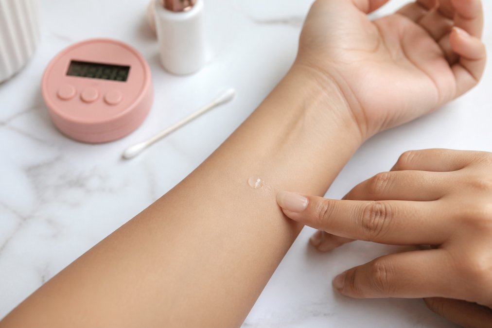 Close-up of a person's inner arm during a patch test, showing a small drop of lash glue applied to clean skin with a timer and cotton swab nearby on a clean surface