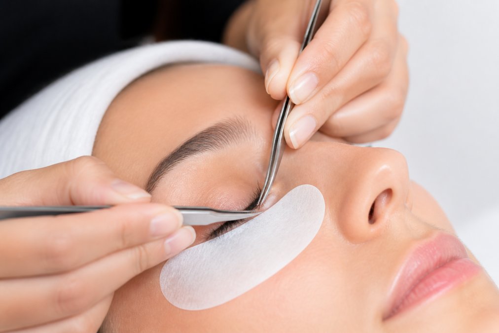 Close-up of a professional lash technician carefully applying ultra-fine lash extensions to a client's delicate lash line under bright clinical lighting, showing the precise attachment point and the contrast between the thin natural lash and the fine extension fiber