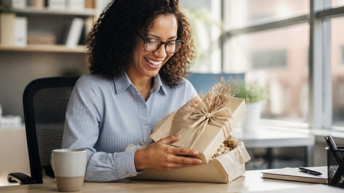 Professional opening a beautifully wrapped handcrafted gift at their office desk with genuine appreciation and a warm smile, natural office lighting streaming through nearby windows