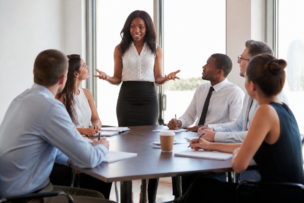 A confident facilitator leading a small group workshop around a table, with participants taking notes and engaging in discussion in a bright, modern training room