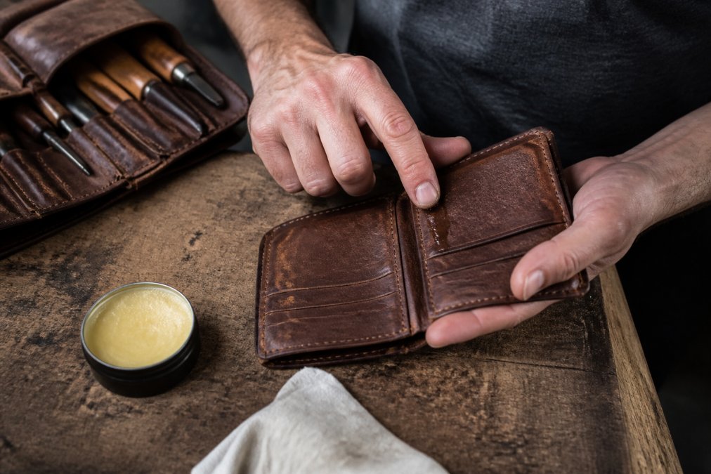 Close-up of a man's hands applying leather conditioner to a well-worn full-grain bifold wallet on a wooden workbench, with a small tin of conditioner and a soft cloth visible nearby